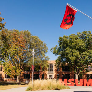 A red flag attached to a brick building in the foreground, with large red letters reading "MSUM" in the background.